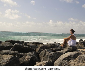 Pretty woman in white dress sitting on rocks watching the ocean