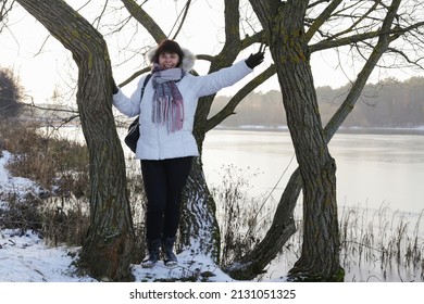 Pretty Woman In White Jacket With Scarf Standing Near Tree In Winter River Background.