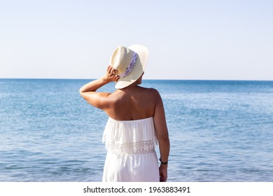 Pretty woman with white off-the-shoulder dress is standing by the water, she has a white hat on her head, she holds it with one hand. She is looking into the distance. Turkey, fame residence, Lara