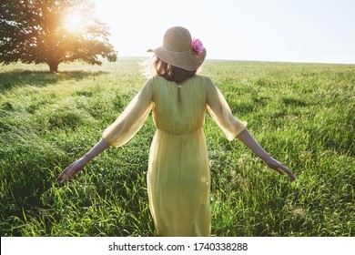 Pretty woman with yellow dress and hat walking in summer field in sunlght