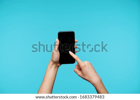 Pretty woman's raised hands with white manicure keeping smartphone and swiping across screen with index finger, posing against blue background