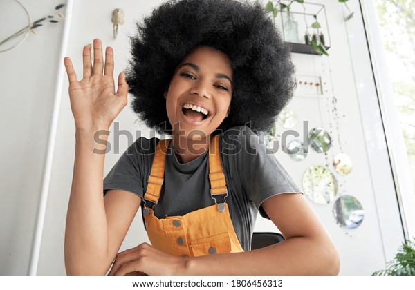 Pretty young African American mixed race\
hipster vlogger woman with Afro hair waving hand looking at webcam\
talking to camera sits at cafe table video calling, recording blog,\
headshot portrait.