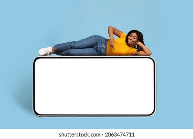 Pretty young african american woman in stylish casual outfit chilling on huge smartphone with empty screen, pointing down and smiling at camera, entertaining mobile app concept, mockup