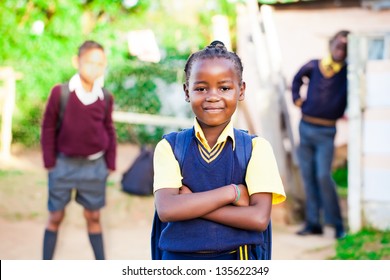 pretty young african girl standing proud in her yellow and blue school uniform with siblings watching over her.