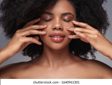 Pretty young African woman closing eyes and touching flawless soft skin on cheeks while representing beauty industry