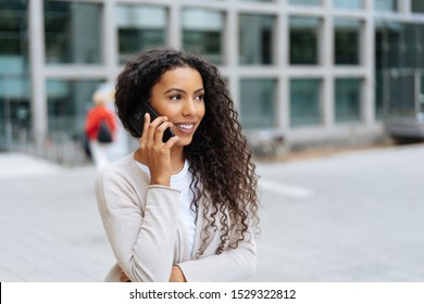 Pretty young African woman with long frizzy hair standing in an urban street chatting on a mobile phone looking aside with a thoughtful smile - Shutterstock ID 1529322812