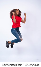 pretty young african woman wearing red top and blue jeans jumping with excitement and happiness, on white background