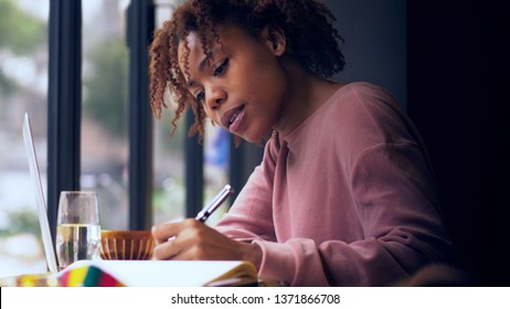pretty young African woman writing indoor Young Black female college student study indoor by the window doing homework