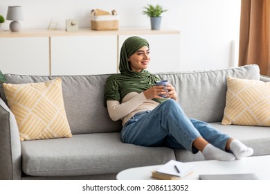 Pretty young Arab lady in hijab sitting on sofa with hot drink, spending peaceful weekend at home, copy space. Beautiful middle Eastern woman in headscarf having coffee break, relaxing on couch