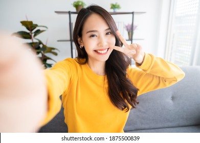 Pretty Young Asian Female With Big Smile Sitting At Living Room. She Having Fun Taking Light Cheerful Selfie On Blurred Background