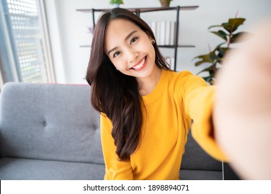 Pretty young asian female with big smile sitting at living room. She having fun taking light cheerful selfie on blurred background