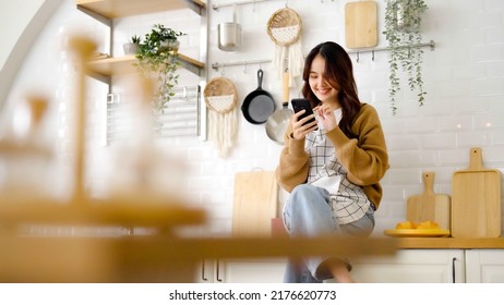 Pretty young asian female smile and sitting on counter kitchen room. She using mobile smart phone having fun video call talking with her friend - Powered by Shutterstock