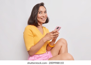 Pretty young Asian woman with bob hairstyle smiles gently has dreamy expression sits against white background holds mobile phone sends text messages wears yelow jumper pink shorts feels happy
