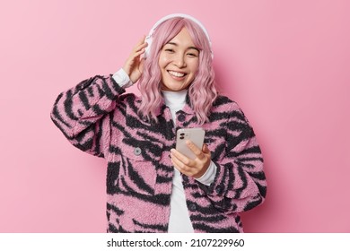 Pretty young Asian woman with dyed hair listens music via stereo headphones holds mobile phone dressed in winter jacket has good mood isolated over pink background. People and hobby concept.