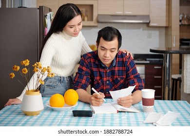 Pretty Young Asian Woman Looking At Boyfriend Checking Utility Bills And Writing Sum Of Expenses In Planner