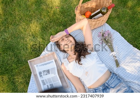 Pretty young asian woman lying on picnic cloth with flower, basket, fruits and newspaper on grass in sunny summer day