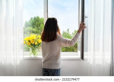 Pretty young Asian woman opening window at home on sunny day, back view - Shutterstock ID 2132179477