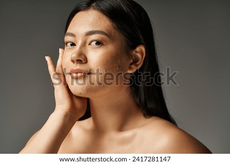 pretty young asian woman with skin issues and bare shoulders looking at camera on grey background