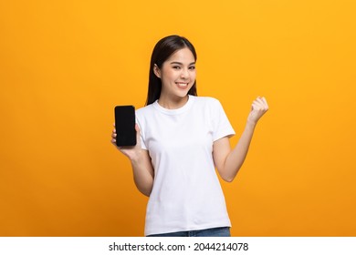 Pretty Young Asian Woman Using Smartphone Standing On Isolated Yellow Background Feeling Happy. Shopping Online Payment With Mobile Phone. Excited And Surprised Female Holding Cellphone.