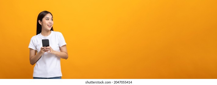 Pretty Young Asian Woman Using Smartphone Standing On Isolated Yellow Background Feeling Happy Looking Blank Space. Shopping Online Payment With Mobile Phone. Excited Female Holding Cellphone.