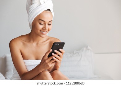 Pretty Young Asian Woman Wrapped In A Shower Towel Using Mobile Phone While Sitting On Bed