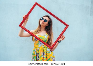 a pretty young attractive girl in bright colorful dress holding a vintage wooden red frame in her hands. the frame space is good for a text message  - Shutterstock ID 1071634304