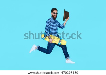 Pretty young bearded man jumping with yellow skateboard against the colorful wall. Hipster in motion on blue background
