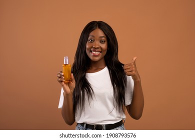 Pretty Young Black Woman With Long Beautiful Hair Holding Bottle With Hair Product, Serum Or Oil, Beige Studio Background, Copy Space. Hair Care, Healthy And Beautiful Hair Concept