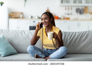 Pretty Young Black Woman With Long Braids In Casual Outfit Having Phone Conversation With Friends Or Lover While Chilling On Couch At Home, Drinking Tea, Looking At Copy Space And Smiling