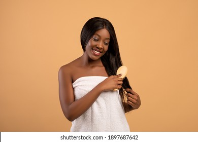 Pretty Young Black Woman Wrapped In Towel Brushing Her Long Beautiful Hair After Shower, Beige Studio Background, Copy Space. Hair Care, Healthy And Beautiful Hair Concept