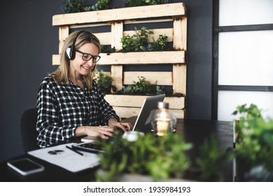 pretty, young blond businesswoman with black and white checked shirt is sitting in a sustainable, ecological office with a headset and is having a web meeting
