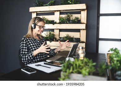 pretty, young blond businesswoman with black and white checked shirt is sitting in a sustainable, ecological office with a headset and is having a web meeting