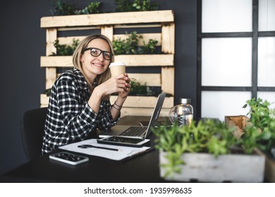 Pretty, Young Blond Businesswoman With A Black And White Checked Shirt Sits In A Sustainable, Ecological Office And Drinks Coffee To Go