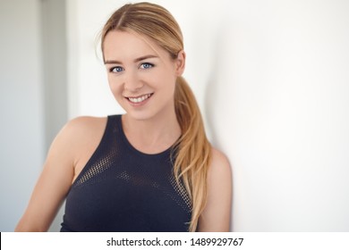 Pretty Young Blond Woman With A Friendly Smile And Her Hair In A Ponytail Leaning On An Interior White Wall Looking At The Camera In A Head And Shoulders Closeup Portrait