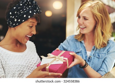 Pretty young blond woman giving an attractive young African American friend a surprise gift gift-wrapped with red paper, a bow and card - Shutterstock ID 324558386