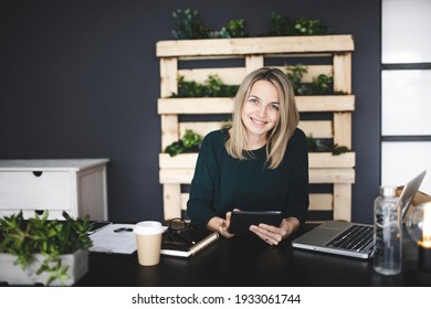 pretty young blond woman is sitting in a modern, sustainable office with lots of green ecological plants and is working on her tablet