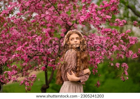 Pretty young blonde girl with long curly hair in vintage dress, apron and bonnet standing in spring park near pink blossom flowers. Tenderness romantic  model posing.