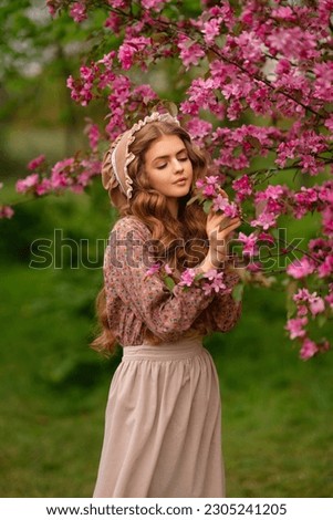 Pretty young blonde girl with long curly hair in vintage dress, apron and bonnet standing in spring park near pink blossom flowers. Tenderness romantic  model posing.