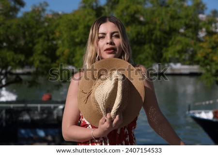 Pretty young blonde woman with straw hat and white dress with red print is having fun doing different poses with the hat close to her face. In the background the river and trees