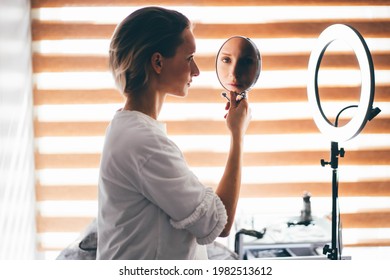 Pretty Young Blonde Woman In White Blouse Looks In Round Mirror Examining Face Sitting On Couch Near Glowing Ring Light In Office Of Cosmetology Clinic.