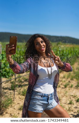 Pretty young Brazilian woman taking a selfie photo with a smartphone outdoors in strong light. Girl smiling expressing energy in good day. Lovely curly woman with good vibes