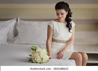 Pretty Young Bride In Simple Wedding Dress With Her Wedding Bouquet. Black-haired Woman With Wedding Hair-style With A Long Tail. Boudoir Morning Of The Bride In Modern Room