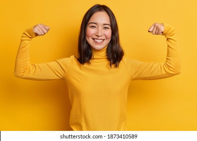 Pretty Young Brunette Asain Woman Raises Arms And Shows Biceps Demonstrates Muscles Feels Proud About Personal Achievements Wears Yellow Turtleneck Stands Indoor. People Strength And Power Concept