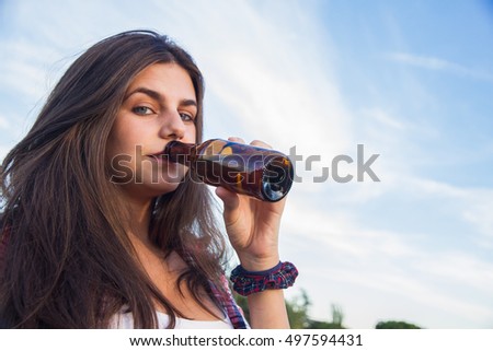 Pretty young brunette with blue eyes drinking from a bottle of beer in the park. She is celebrating the holidays and she is happy. Background is blue sky. 