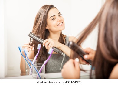 Pretty young brunette straightening her hair in the bathroom using a flat iron and smiling - Powered by Shutterstock