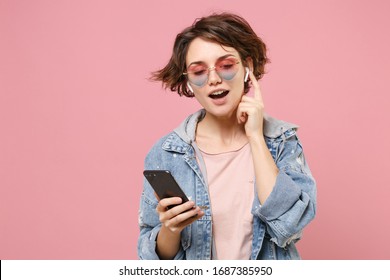 Pretty Young Brunette Woman In Casual Denim Jacket, Eyeglasses Posing Isolated On Pastel Pink Background. People Lifestyle Concept. Mock Up Copy Space. Listen Music With Air Pods, Hold Mobile Phone