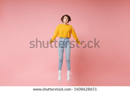 Pretty young brunette woman girl in yellow sweater posing isolated on pastel pink background in studio. People lifestyle concept. Mock up copy space. Having fun spreading hands rising hands, jumping