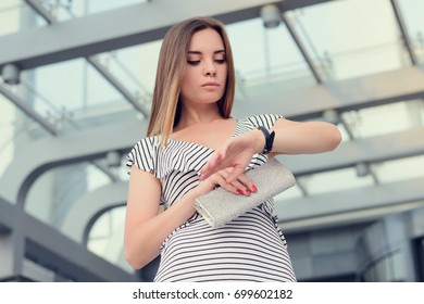 Pretty young businesslady in striped dress with purse in  hand checking the time on her watch and waiting for colleague near office building