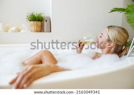 Pretty young Caucasian female relaxing in bath with bubbles and sipping white wine.