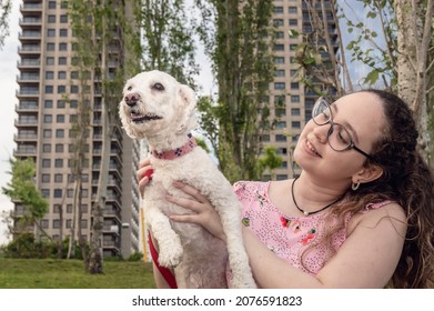 Pretty Young Caucasian Girl With Curls And Glasses Sitting In A Beautiful Urban Park Outside Her Building, Playing With Her White Poodle Dog, Lifestyle Concept.